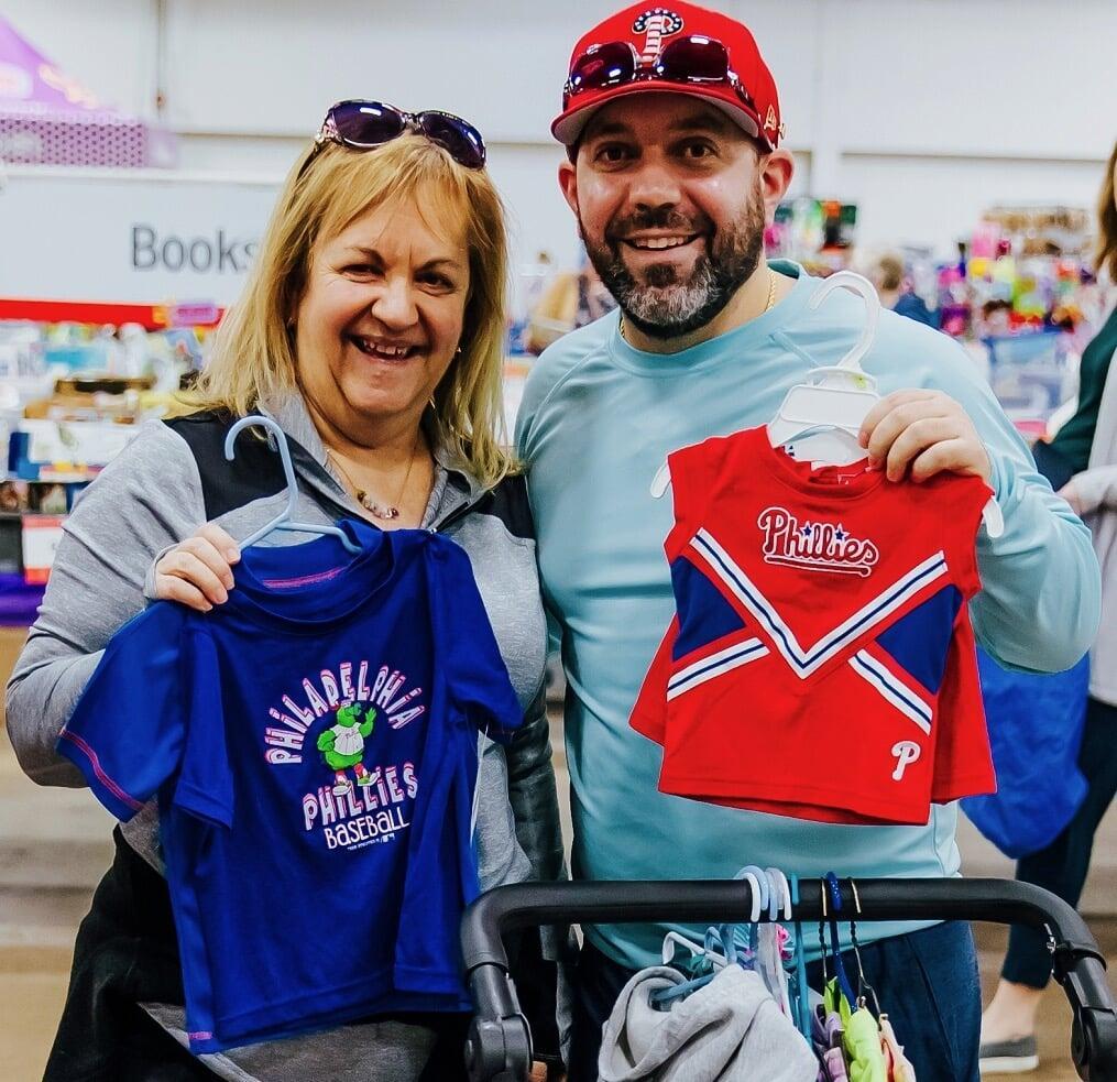 Smiling mom and dad, both holding clothing on hangers.  Dad has baby in a front pack Baby Bjorn carrier.