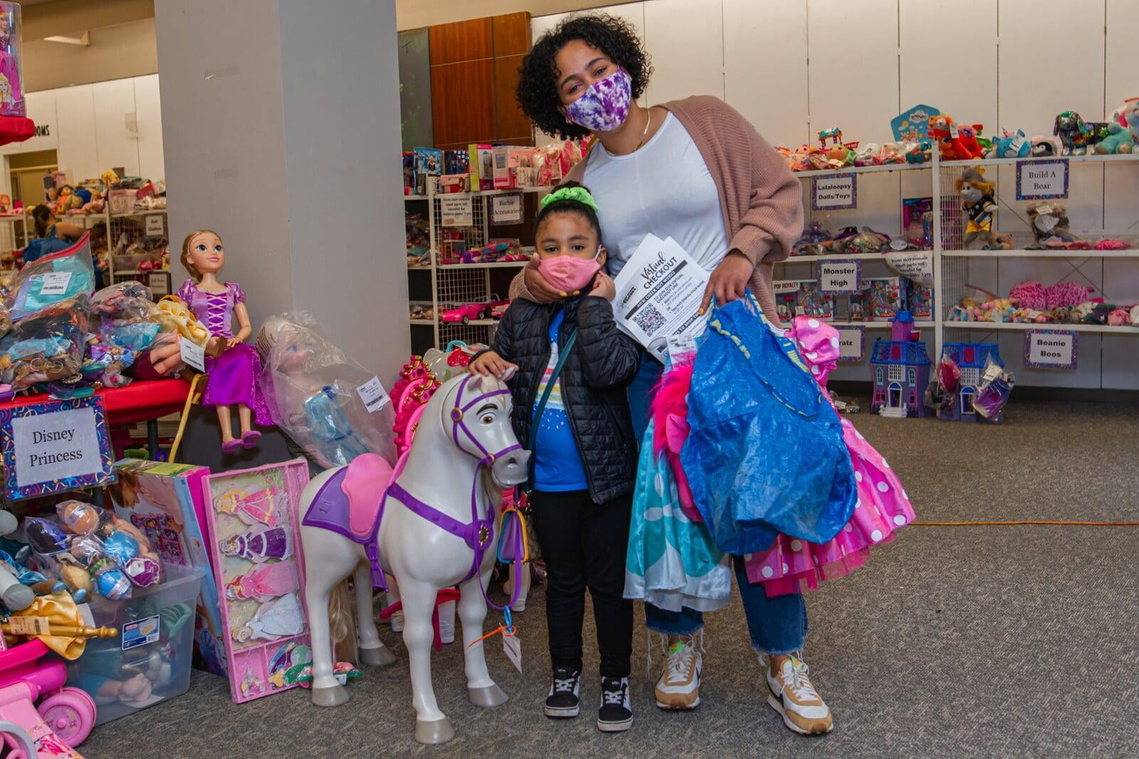 A mom standing with her arm around her daughter & the daughter's hand is on a toy horse.  Mom is holding a JBF shopping bag and clothing.